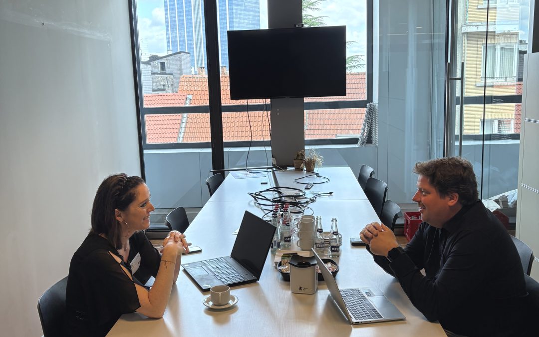 Fabienne Colling, Vorsitzende der Ecolo Fraktion des PDG, sitzt gegenüber von Jeremie Vaneeckhout der Groen-Fraktion im Flämischen Parlament an einem Schreibtisch. Im Hintergrund sieht man ein Fenster mit Blick auf Gebäude.
